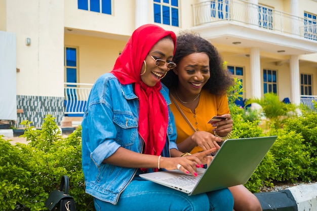 Zwei hübsche afrikanische Damen, die ihre Kreditkarte und ihren Laptop benutzen, um online einzukaufen.