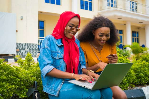 Zwei hübsche afrikanische Damen, die ihre Kreditkarte und ihren Laptop benutzen, um online einzukaufen.