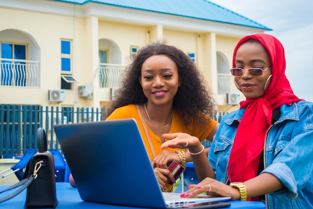 Zwei hübsche afrikanische Damen, die ihre Kreditkarte und ihren Laptop benutzen, um online einzukaufen.