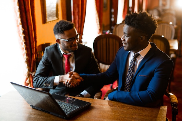 zwei hübsche afrikanische Männer drinnen mit Laptop im Handschlag