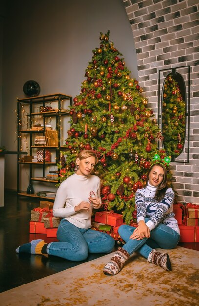 Zwei hübsche Frauen schließen unter dem festlichen Tannenbaum drinnen mit den Kisten der Geschenke. Weihnachtsfoto