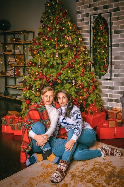 Zwei hübsche Frauen schließen unter dem festlichen Tannenbaum drinnen mit den Kisten der Geschenke. Weihnachtsfoto