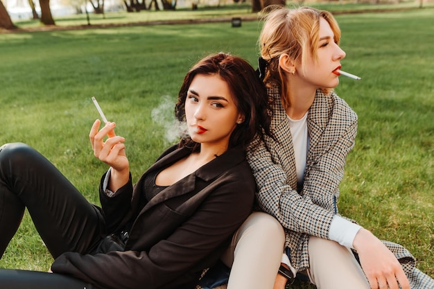 Zwei hübsche Freundinnen sitzen auf Gras und rauchen eine Zigarette. Ein paar schwule lesbische Mädchen umarmen sich