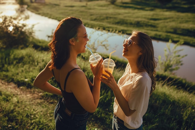 Zwei hübsche Freundinnen trinken Cocktail, gekleidet in Schwarz-Weiß-T-Shirt, Spaß im Sommer, bei Sonnenuntergang, positiver Gesichtsausdruck, im Freien