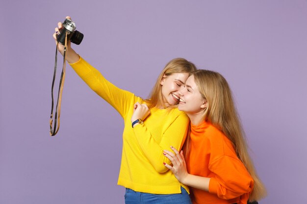 Zwei hübsche junge blonde Zwillingsschwestern Mädchen in bunten Kleidern machen Selfie-Aufnahme auf Retro-Vintage-Fotokamera isoliert auf violettblauer Wand Menschen-Familien-Lifestyle-Konzept.