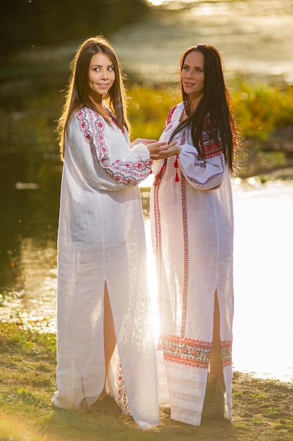 Zwei hübsche junge Damen tragen traditionelle ukrainische Kleidung und Blumenkranz Spaziergang im Weizenfeld schönes ethnisches Mädchen in handgefertigter Blumenkrone bewundern die Natur Sonnenuntergang