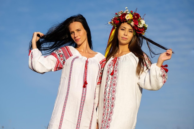 Zwei hübsche junge Damen tragen traditionelle ukrainische Kleidung und Blumenkranz Spaziergang im Weizenfeld schönes ethnisches Mädchen in handgefertigter Blumenkrone bewundern die Natur Sonnenuntergang