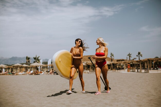 Zwei hübsche junge Frauen mit Paddle Board am Strand an einem Sommertag