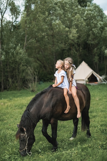 Zwei hübsche kleine Mädchen in lässiger Boho-Kleidung galoppieren auf einem schönen dunklen Pferd im Freien