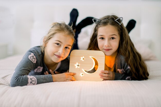 Zwei hübsche kleine Schwestern, die zu Hause auf dem Bett liegen und ihre Zeit genießen, mit hölzerner Nachtlampe mit Mond- und Sternenbild spielend.