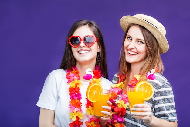 Zwei hübsche lächelnde Mädchen mit Lei am Hals halten Orangengetränke vor violettem Hintergrund