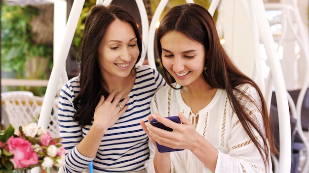 Zwei hübsche Mädchen lachen beim Betrachten von Fotos am Telefon, die in einem Café sitzen.