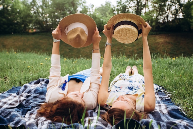 Zwei hübsche Mädchen liegen auf einem blauen Plaid draußen im Park, Sommerpicknick. Die Mädchen der Freundinnen haben Spaß, lachen, lächeln und toben. Nahaufnahmeporträt von zwei jungen Frauen in Kleidern und Strohhüten.
