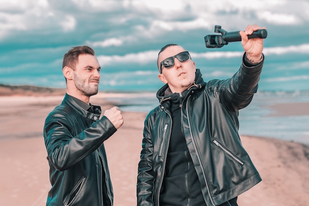 Zwei hübsche männliche Freunde, die Selfie unter Verwendung der Aktionskamera mit kardanischem Stabilisator am Strand machen.