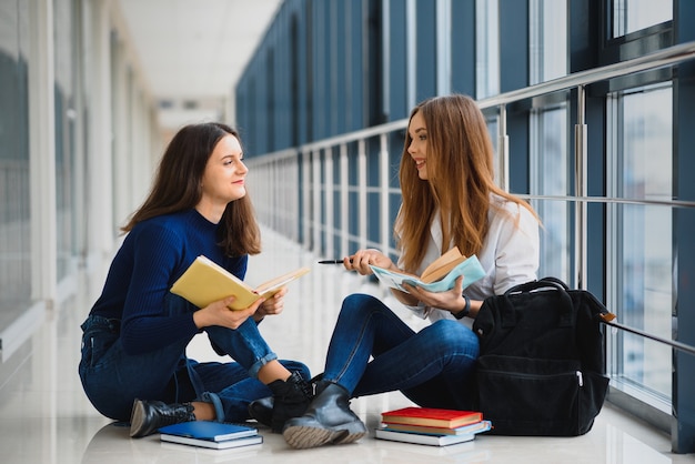 Zwei hübsche Studentinnen mit Büchern, die auf dem Boden im Flur der Universität sitzen