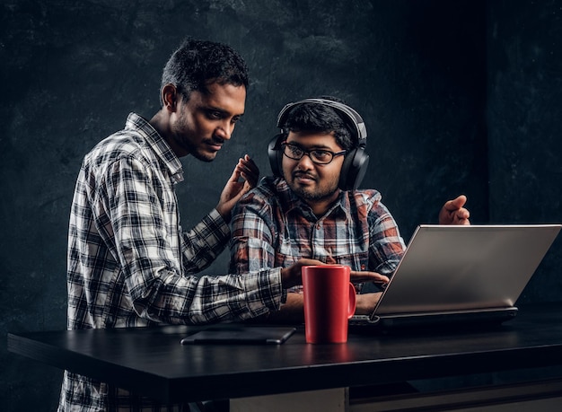 Zwei indische Studentenfreunde, die gemeinsam an einem neuen Projekt arbeiten, sitzen mit einem Laptop an einer dunklen, strukturierten Wand am Tisch.