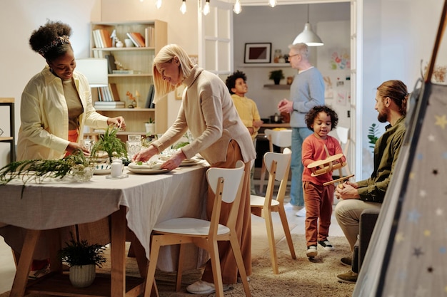 Zwei interkulturelle Frauen bedienen gemeinsam mit anderen Familienmitgliedern den Tisch zum Abendessen