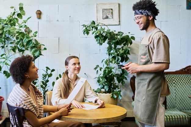 Zwei interkulturelle Mädchen mit Menü, die einen lächelnden Kellner in Arbeitsbekleidung ansehen