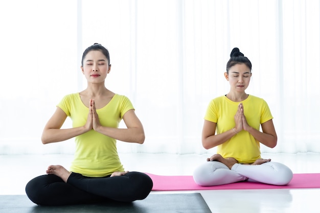 Zwei junge asiatische Frauen trainieren Yoga im gelben Kleid oder posieren mit einem Trainer und praktizieren Meditation, Wellness-Lifestyle und Gesundheits-Fitness-Konzept in einem Fitnessstudio.