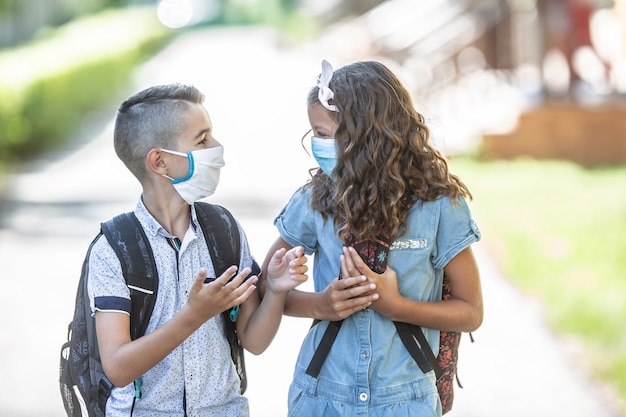 Zwei junge befreundete Klassenkameraden mit Gesichtsmasken unterhalten sich während der Covid-19-Quarantäne auf dem Weg zur Schule.