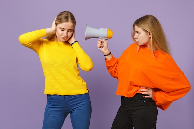 Zwei junge blonde Zwillingsschwestern Mädchen in bunten Kleidern, die Ohren mit Händen bedecken, schreien auf Megaphon einzeln auf pastellvioletter blauer Wand. Menschen-Familien-Lifestyle-Konzept. .