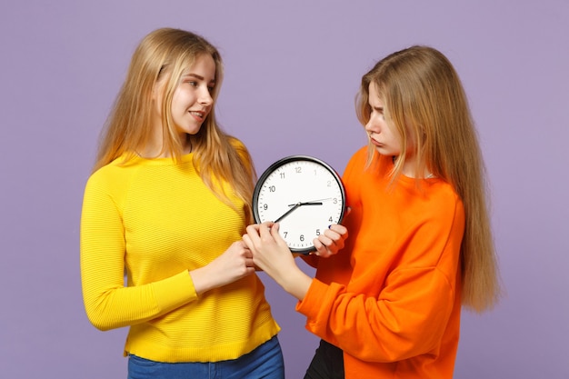 Zwei junge blonde Zwillingsschwestern Mädchen in lebendigen bunten Kleidern mit runder Uhr, bewegliche Pfeile einzeln auf pastellvioletter blauer Wand. Menschen-Familien-Lifestyle-Konzept.
