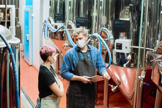 Zwei junge Brauer einer großen modernen Bierproduktionsfabrik beraten über einige Arbeitsmomente zwischen Stahltanks