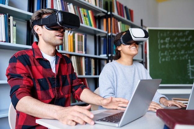 Zwei junge College-Studenten in VR-Headsets beobachten die virtuelle Präsentation, während sie in der Bibliothek oder im Klassenzimmer sitzen