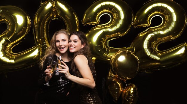 Zwei junge Damen trinken Champagner. Bild von Mädchen mit Luftballons auf schwarzem Hintergrund, die Spaß an Weihnachten oder Silvester 2022 haben.