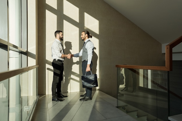 Zwei junge elegante Geschäftspartner Händeschütteln, nachdem sie einen neuen Vertrag ausgehandelt und unterschrieben haben, während sie an der Wand standen