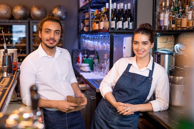 Zwei junge erholsame Arbeiter des Cafés, die nebeneinander vor der Kamera auf Hintergrund des Regals mit Weinsortiment stehen