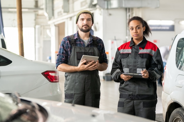 Zwei junge ernsthafte Arbeiter des Autowartungszentrums in Uniform stehen zwischen Autos vor der Kamera, während einer von ihnen ein Tablet benutzt