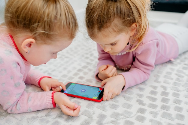 Zwei junge fokussierte Kinder, die ein Smartphone ohne Namen spielen, das auf dem Boden des Wohnzimmers liegt. Kleine Kinder und Technik, Schwestern spielen mit einem Handy, schauen sich Videos an oder spielen Spiele