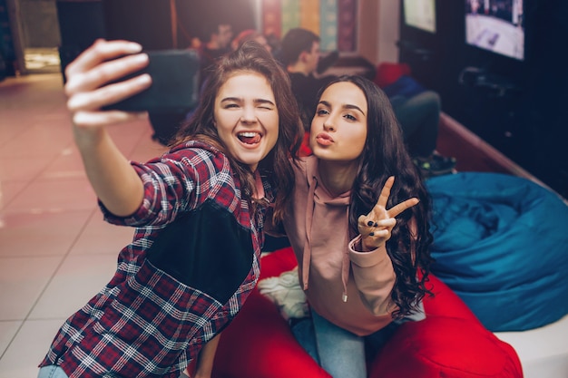 Zwei junge Frauen, die Selfie auf Telefonkamera nehmen und im Spielzimmer posieren. Sie schimmern und zeigen Finger. Fröhliche Models lächeln.