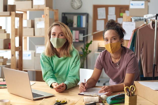 Zwei junge Frauen in Schutzmasken arbeiten zusammen am Tisch mit Laptop in der Werkstatt