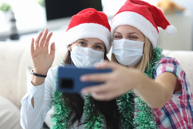 Zwei junge Frauen in Weihnachtsmützen und Schutzmasken auf Gesichtern halten Telefon zu Hause Porträt