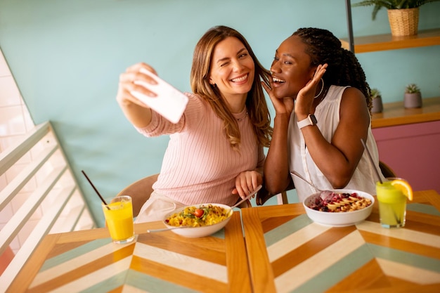 Zwei junge Frauen, Kaukasier und Schwarze, die im Café ein Selfie mit dem Handy machen