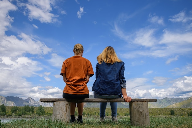 Zwei junge frauen sitzen auf holzbank, rücken zur kamera gegen schöne landschaft. freunde reisen, bewundern sie die herrliche aussicht auf die bergkette unter blauem himmel an einem sonnigen tag.