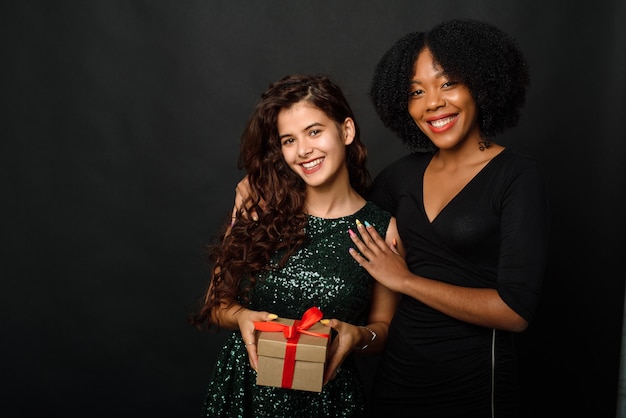 Zwei junge Frauen verschiedener Nationalitäten umarmen sich und halten ein Geschenk für das neue Jahr und Weihnachten auf einem schwarzen Hintergrund.