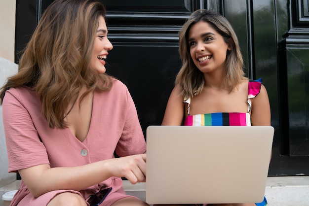 Zwei junge Freunde, die zusammen einen Laptop benutzen, während sie draußen auf der Straße sitzen. Stadt- und Technologiekonzept.