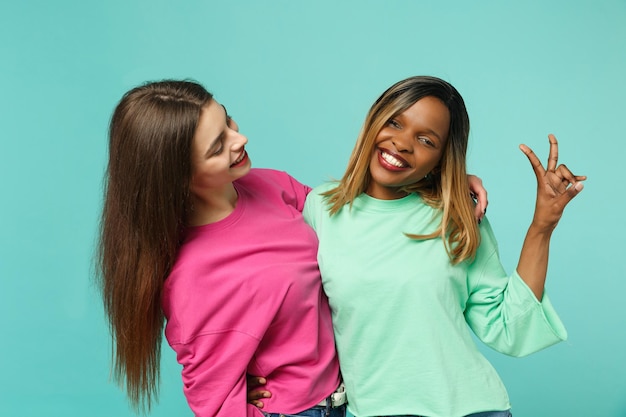Zwei junge Freundinnen europäisch und afroamerikanisch in rosa grüner Kleidung stehen isoliert auf blauem türkisfarbenem Wandhintergrund, Studioporträt. People-Lifestyle-Konzept. Mock-up-Kopienbereich.