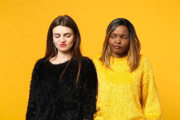 Zwei junge Freundinnen europäisch und afroamerikanisch in schwarz-gelber Kleidung stehen isoliert auf leuchtend orangefarbenem Wandhintergrund, Studioporträt. People-Lifestyle-Konzept. Mock-up-Kopienbereich.