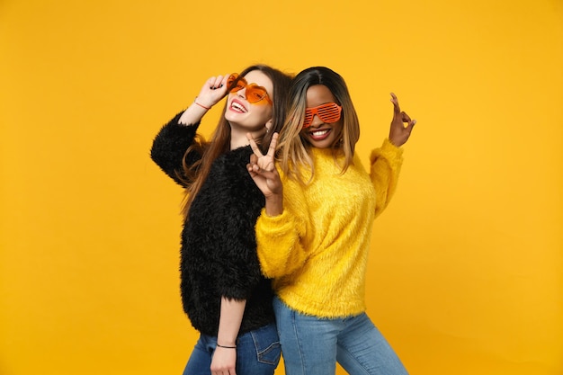 Zwei junge Freundinnen europäisch und afroamerikanisch in schwarz-gelber Kleidung stehen isoliert auf leuchtend orangefarbenem Wandhintergrund, Studioporträt. People-Lifestyle-Konzept. Mock-up-Kopienbereich.