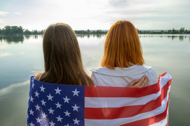Zwei junge Freundinnen mit USA-Nationalflagge auf ihren Schultern, die zusammen draußen am Seeufer stehen.