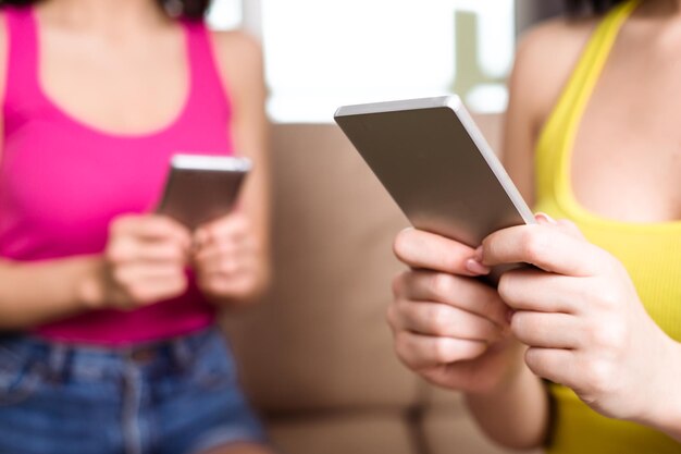 Zwei junge Freundinnen sitzen zu Hause auf einem Sofa und schreiben Nachrichten am Telefon oder nutzen das Internet