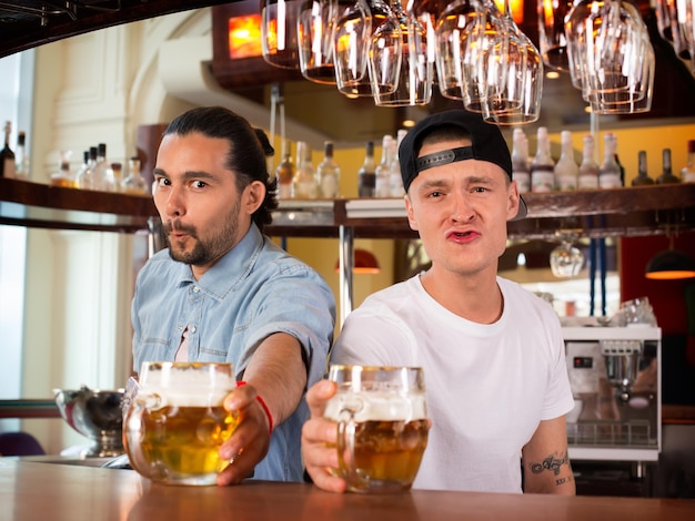 Zwei junge, fröhliche Barkeeper schauen in die Kamera, machen lustige Gesichter und bieten den Kunden Bier an. Fröhliche Barkeeper, die bei der Arbeit albern sind, einen schönen Tag haben und froh sind, Menschen zu dienen.