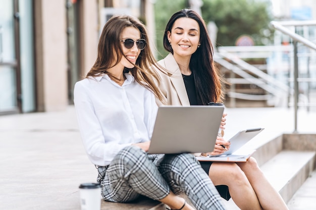 Zwei junge Geschäftsdame mit Tablet und Laptop, die draußen in der Nähe des modernen Geschäftszentrums sitzen.