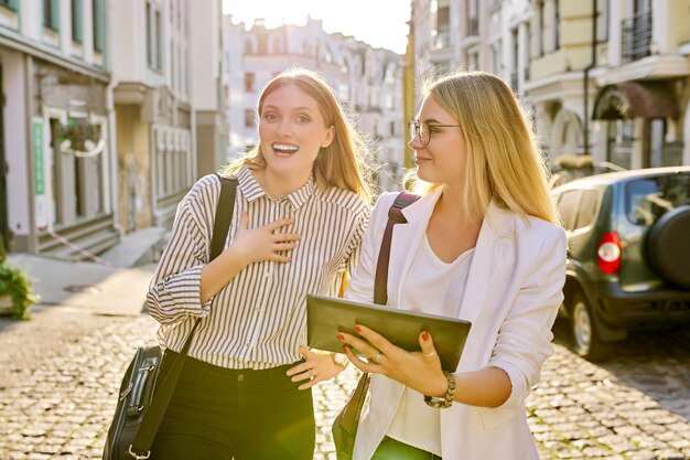 Zwei junge Geschäftsfrauen gehen entlang der Stadtstraße und unterhalten sich und schauen auf den Bildschirm eines digitalen Tablets