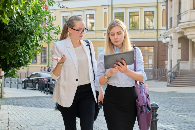 Zwei junge Geschäftsfrauen gehen entlang der Stadtstraße, unterhalten sich und schauen auf den Bildschirm eines digitalen Tablets. Business, Menschen, Technik, Teamwork, Kommunikationskonzept