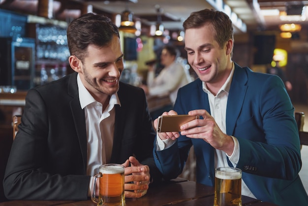 Zwei junge Geschäftsleute in Anzügen lächeln mit einem Handy und trinken Bier, während sie in der Kneipe sitzen